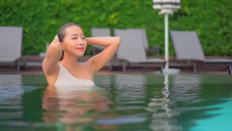 Sexy-asian-woman-standing-in-a-swimming-pool-water-half-submerged,-closing-her-eyes-looking-up-and-touching-her-wet-hair-enjoying-her-summer-holidays