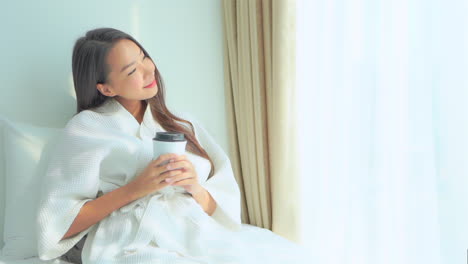 A-beautiful-young-woman-relaxes-on-a-hotel-suite-bed-in-her-robe-while-holding-a-cup-of-coffee-in-her-hands