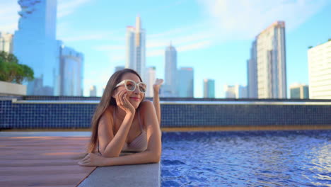 Sexy-exotic-asian-female-in-swimsuit-enjoying-by-rooftop-pool-with-a-modern-city-buildings-in-background,-full-frame-slow-motion