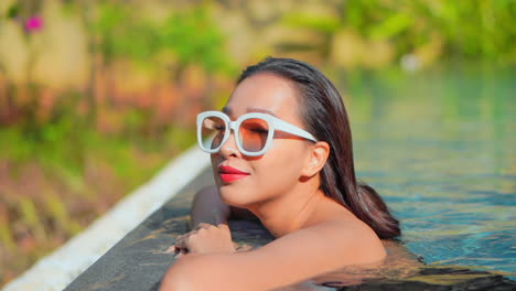 Side-view-of-a-female-tourist-wearing-big-white-sun-glasses,-leaning-on-the-edge-of-an-outdoor-swimming-pool-in-some-tropical-destination,-putting-her-head-on-the-edge-and-smiling,-looking-happy