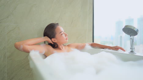 A-beautiful-young-woman-lounges-in-a-bubble-bath-as-she-takes-in-the-modern-world-through-her-window-from-her-high-rise-building