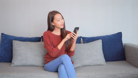 Asian-woman-playing-a-mobile-game-on-her-smartphone-touching-screen-with-one-finger-sitting-on-comfortable-coach-with-many-pillows-at-home