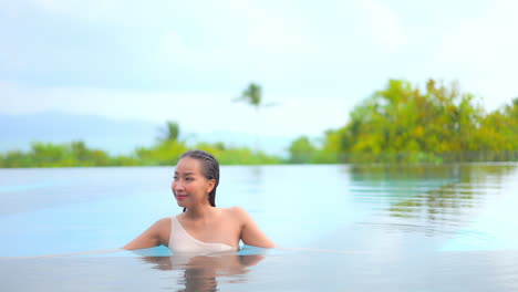 Asian-woman-standing-half-submerged-in-the-swimming-pool-water,-looking-around-,-with-tropical-out-of-focus-background-behind-her