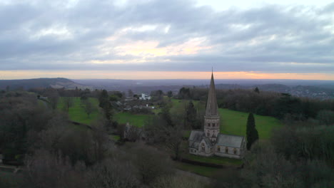 An-aerial-shot-flying-past-an-old-English-church-on-a-hill-in-Ranmore-common,-in-Dorking-at-sunrise