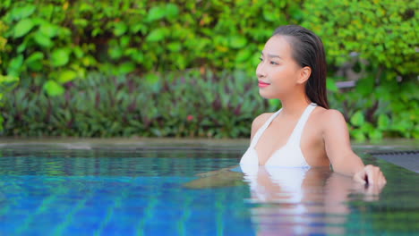 Portrait-of-a-happy-smiling-brunette-woman-in-white-swimsuit-relaxing-in-swimming-pool-and-looking-in-aside-in-Thailand-resort