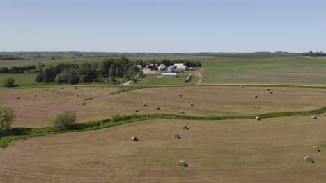 Low-Establishing-Aerial-Shot-of-Hay-Bales-in-Empty-Field-with-Farm-in-Background