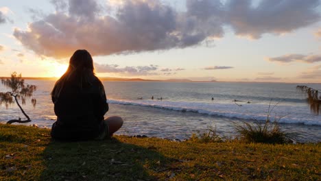 Blick-Hinter-Eine-Frau,-Die-An-Der-Küste-Sitzt-Und-Surfer-In-Der-Lagunenbucht-Mit-Sonnenbeschienenem-Hintergrund-Beobachtet