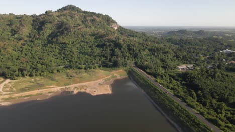 4k-luftaufnahmen-Des-Khlong-Bod-reservoirs-Mit-Damm-Und-Straße,-Die-Zum-Hügel-Mit-Grünen-Tropischen-Bäumen-In-Nakhon-Nayok,-Thailand-Führen