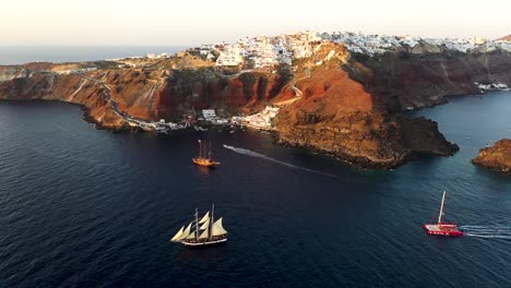 Aerial-drone-view-video-of-beautiful-sail-boats-cruising-in-the-deep-dark-blue-Aegean-sea-during-sunset,-Santorini,-Cyclades,-Greec