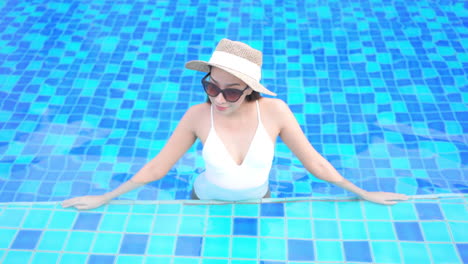 Asian-Woman-Resting-Inside-Swimming-Pool,-Wearing-White-Swimsuit,-Sunhat-and-Sunglasses-and-Leaning-on-Pool's-Edge---High-Angle-Front-View