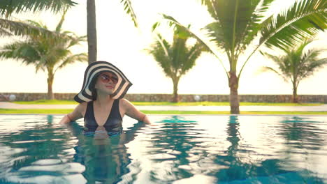 Smiling-Happy-Asian-Lady-in-Large-Black-and-White-Sunhat-Inside-Swimming-Pool-Enjoying-Tropical-Vacation-Leaning-on-Pool's-Border-At-Sunset
