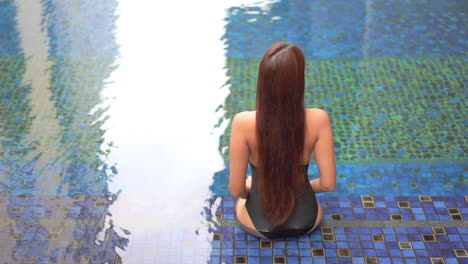 Back-view-of-the-fit-Asian-woman-in-black-swimsuit-sitting-in-shallow-water-on-the-edge-of-a-swimming-pool,-elevated-point-of-view-slow-motion