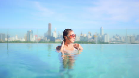 Woman-smooth-wet-hair-in-rooftop-swimming-pool-with-view-of-Bangkok-city-skyline,-slow-motion-copy-space