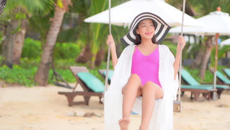 Asiatische-Touristin-Lächelt,-Während-Sie-An-Einem-Sonnigen-Tag-Auf-Einer-Schaukel-Am-Tropischen-Strand-Schwingt-Und-Einen-Rosa-Badeanzug-Und-Einen-Großen-Gestreiften-Hut-Trägt