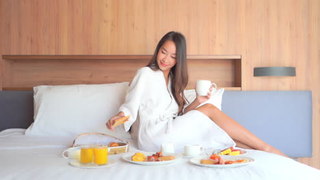 Beautiful-Asian-Girl-in-Bathrobe-Drinking-Coffee-with-Toast-and-Having-Breakfast-in-Bed-in-Hotel-Room,-Plates-with-Fruits-and-Juices-on-a-Made-up-Bed,-slow-motion