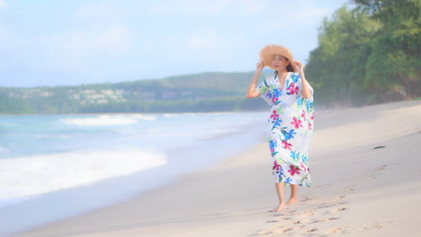 Slow-motion-of-Asian-lady-walking-along-the-beach-in-Thailand-wearing-a-long-sundress,-she-is-holding-her-straw-hat-on-windy-day,-slow-motion-following-show-side-view