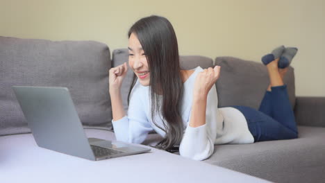 Happy-Asian-young-woman-lying-on-the-sofa-at-home-typing-on-laptop-receive-great-news-celebrating-a-moment-of-victory,-personal-achievement,-got-hired,-high-school-scholarship-admission-concept