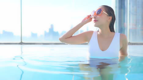 Woman-in-a-rooftop-swimming-pool,-resting-leaning-on-elbows-and-looks-out-over-modern-city-skyline,-Title-space