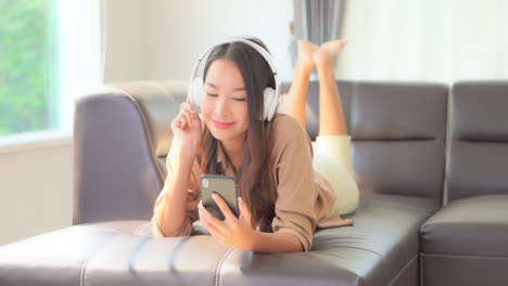 A-young-woman-playfully-kicks-her-feet-while-lying-on-the-couch-as-she-listens-to-music-through-her-smartphone-and-headphones