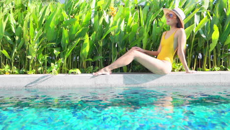 A-full-body-profile-a-young-attractive-woman-on-a-one-piece-bathing-suit-and-sun-hat-sitting-on-the-edge-of-a-swimming-pool-taking-in-the-sun