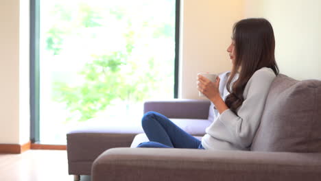 Thai-woman-sitting-in-cozy-sofa-and-drink-hot-coffee-in-the-morning-on-weekend---side-view-slow-motion