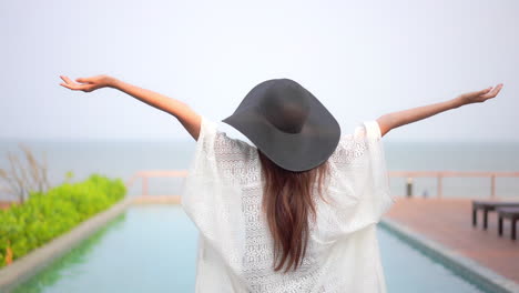 Back-of-Asian-Woman-Standing-by-the-Swimming-Infinity-Pool-in-Summer-Wearing-a-Sundress-and-Black-Hat-Raising-Hands-Up,-Vacation-template