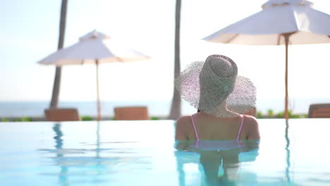 Rückseite-Einer-Schönen-Frau-Mit-Sonnenhut,-Die-Im-Wasser-Eines-Swimmingpools-In-Einer-Tropischen-Insel-Resort-Lounge-Mit-Sonnenschirmen-Und-Liegestühlen-Im-Hintergrund-Steht,-Zeitlupen-Handgerät,-Urlaubsvorlage