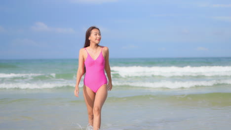Sexy-young-Asian-woman-wearing-pink-monokini-swimwear-walks-on-sea-shore-towards-camera-with-ocean-waves-in-background