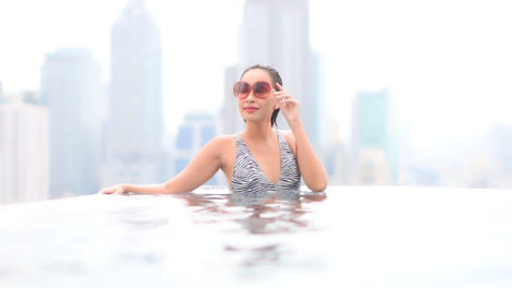 Sensual-slim-Asian-Thai-woman-with-vintage-sunglasses-relaxing-in-luxurious-infinity-pool-with-incredible-view-of-Bangkok-cityscape-in-background