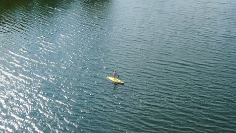Person-paddling-on-yellow-board-on-blue-water,-aerial-orbit-view