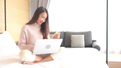 Asian-woman-with-a-beautiful-smile-is-using-her-credit-card-to-make-an-online-payment-using-a-laptop-computer,-money-transfer,-female-sitting-cross-legged-on-the-bed-and-put-in-credit-card-info