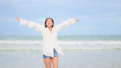 Woman-with-white-shirt-and-shorts-standing-on-seashore-turns-on-herself-raises-arms-up-to-blue-sky
