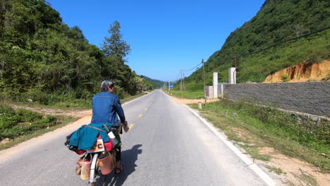 A-cyclist-in-blue-shirt-riding-his-cycle-loaded-with-luggage-on-a-mountain-road-in-Cao-Bang-province,-Vietnam