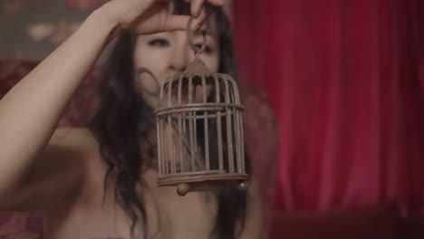 Alternative-girl-with-black-hair-sitting-in-a-big-red-armchair-in-a-gothic-room-holding-a-small-bird-cage-with-a-key
