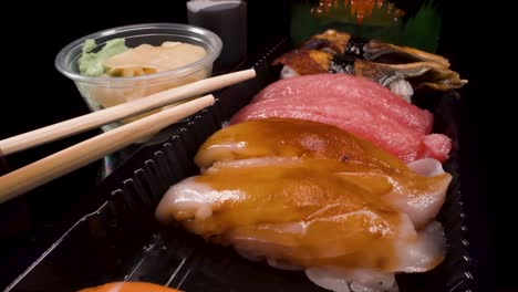 Makroansicht,-Die-über-Take-out-Sushi-Boxen-Auf-Elegantem-Schwarzem-Hintergrund-Fliegt-Und-Nigiri-Sushi,-Lachs,-Thunfisch,-Unagi,-Ikura-Aufnimmt