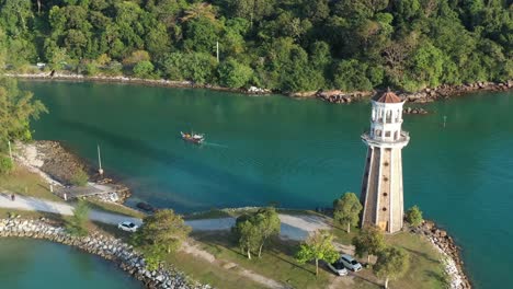 Cinematic-shot-drone-fly-around-perdana-quay-light-house,-following-a-fishing-boat-sailing-on-waterway-toward-telaga-harbor-marina-surrounded-by-beautiful-nature-at-langkawi-island,-kedah,-malaysia