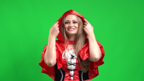 A-beautiful-girl,-wearing-a-Little-Red-Riding-Hood-costume,-in-front-of-a-green-screen,-smiling-while-looking-at-the-camera-and-covering-her-head-with-the-hoodie