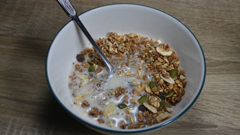 Zoom-out-of-Granola-in-a-bowl-with-a-spoon-and-Almond-Milk-just-poured-in-making-bubbles,-seeds,-dried-fruits,-healthy-breakfast