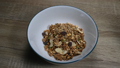 Granola-in-a-bowl-then-a-spoon-is-being-placed-inside-the-bowl-digging-into-the-granola-made-of-seeds,-dried-fruits,-nuts,-raisins,-healthy-breakfast