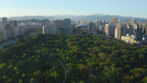 Sobrevuelo-Aéreo-épico-Parque-Daan-Rural-Con-árboles-Verdes-Rodeados-Por-Un-Horizonte-Con-Edificios-Rascacielos-Y-Silueta-De-Montañas-En-El-Fondo---Ciudad-De-Taipei,-Taiwán-Durante-El-Día-Y-El-Sol