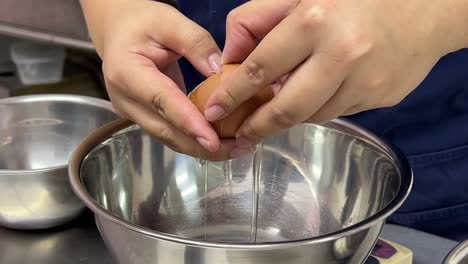 Pastry-chef-cracking-egg,-separating-egg-white-and-egg-yolk-into-different-stainless-steel-mixing-bowl,-commercial-kitchen-bakery-setting
