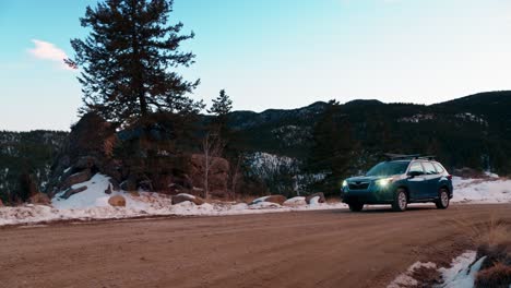 Stunning-tracking-shot-of-a-blue-SUV-driving-along-a-snowy-dirt-mountain-road-during-sunset