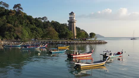 Schöne-Luftaufnahme-Des-Sonnenuntergangs-Von-Perdana-Quay-Light-House-Mit-Traditionellen-Fischerbooten,-Die-Am-Ufer-Festgemacht-Sind,-Mit-Zwei-Segelbooten,-Die-Aus-Dem-Hafen-Von-Telaga-Auf-Der-Insel-Langkawi,-Kedah,-Malaysia-Segeln