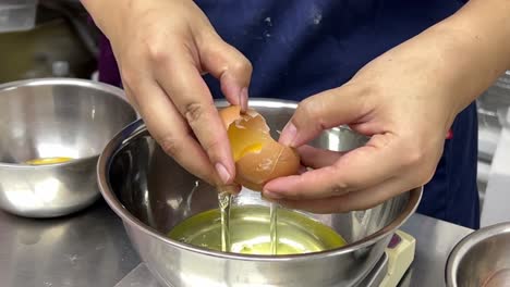 Delicious-dessert-in-making,-pastry-chef-cracking-egg,-separating-egg-white-and-egg-yolk-into-different-stainless-steel-mixing-bowl,-commercial-restaurant-setting
