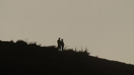 Two-Unrecognizable-People-in-Silhouette-Standing-on-Hill-Slope,-Sunset
