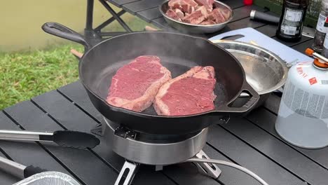 Two-cuts-of-steak-sizzling-on-hot-cast-iron-frying-pan-at-outdoor-camping-site-environment-during-daytime,-close-up-shot