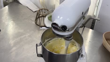 Hand-holding-a-bowl-of-eggs,-slowly-pouring-and-adding-into-the-cake-mixtures,-mixing-in-the-machine-mixer