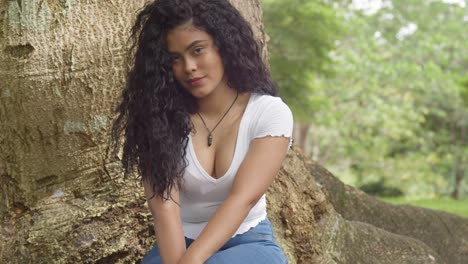 Curly-hair-hispanic-girl-sits-a-the-base-of-a-large-tree-trunk-on-a-sunny-day