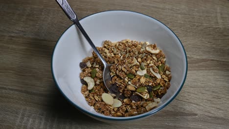 Granola-in-a-bowl-a-spoon-digs-in-and-Almond-Milk-being-poured-in-making-bubbles,-seeds,-dried-fruits,-raisins,-healthy-breakfast