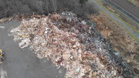 Wide-view-aerial-flying-at-low-altitude-towards-huge-dumpsite-pile-filled-with-tons-of-wooden-and-bulky-waste-made-up-of-euro-pallets-while-tractor-shovels-sawdust